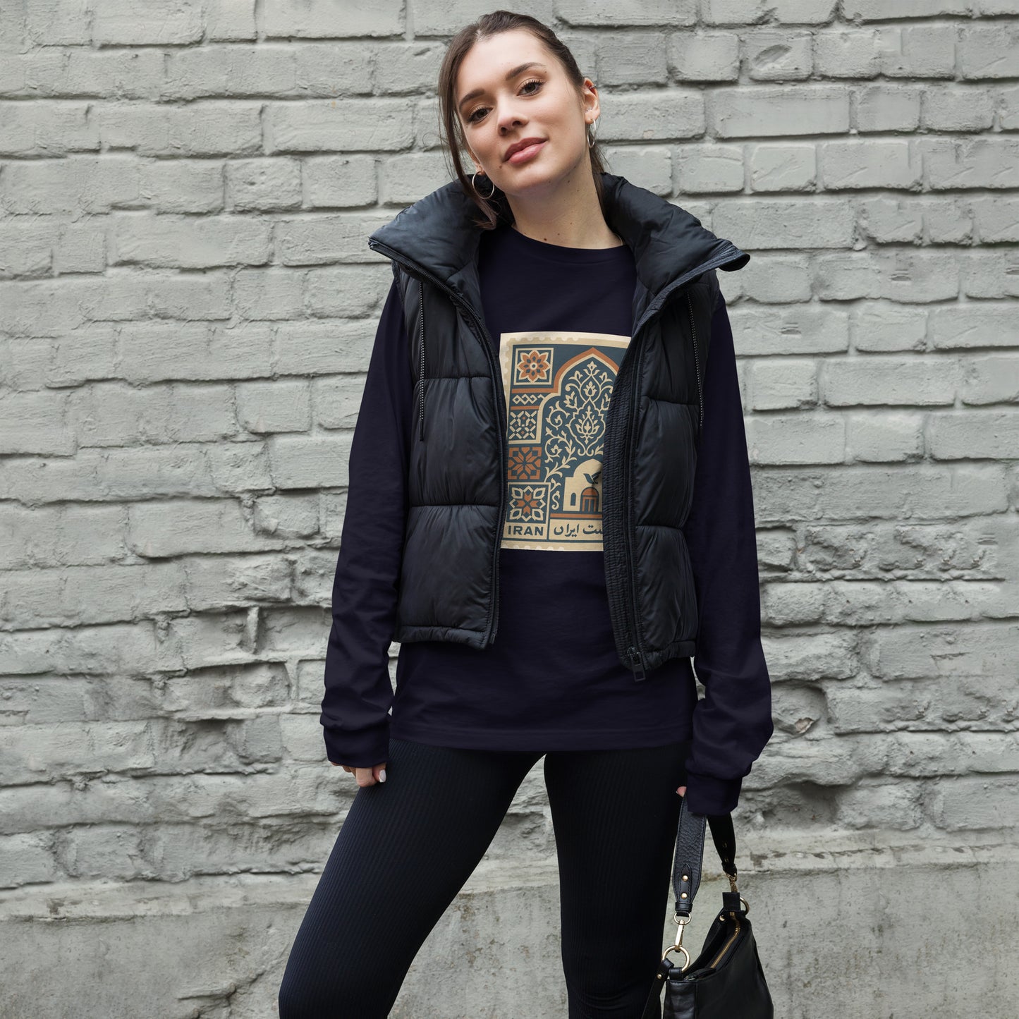 Woman wearing a dark jacket and black pants with a graphic t-shirt against a brick wall.
