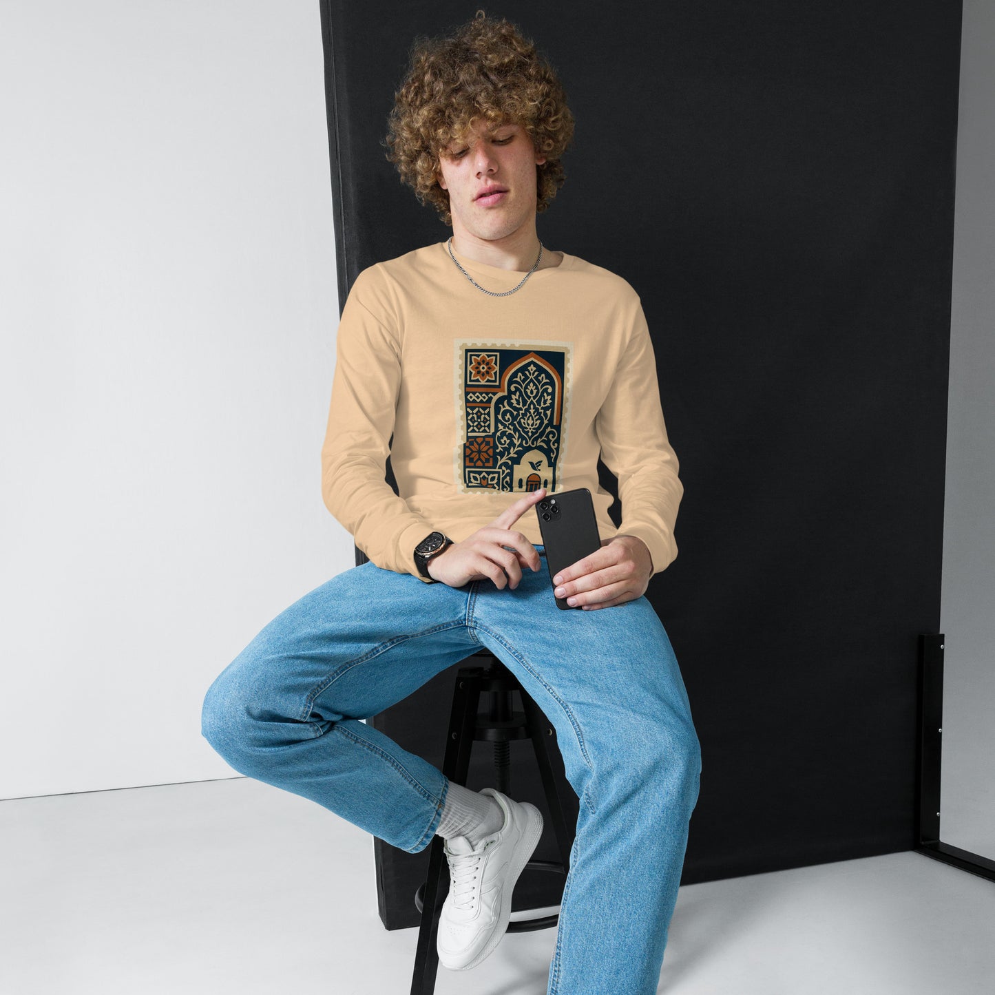 Person wearing a beige sweatshirt with a graphic design, sitting against a black backdrop.