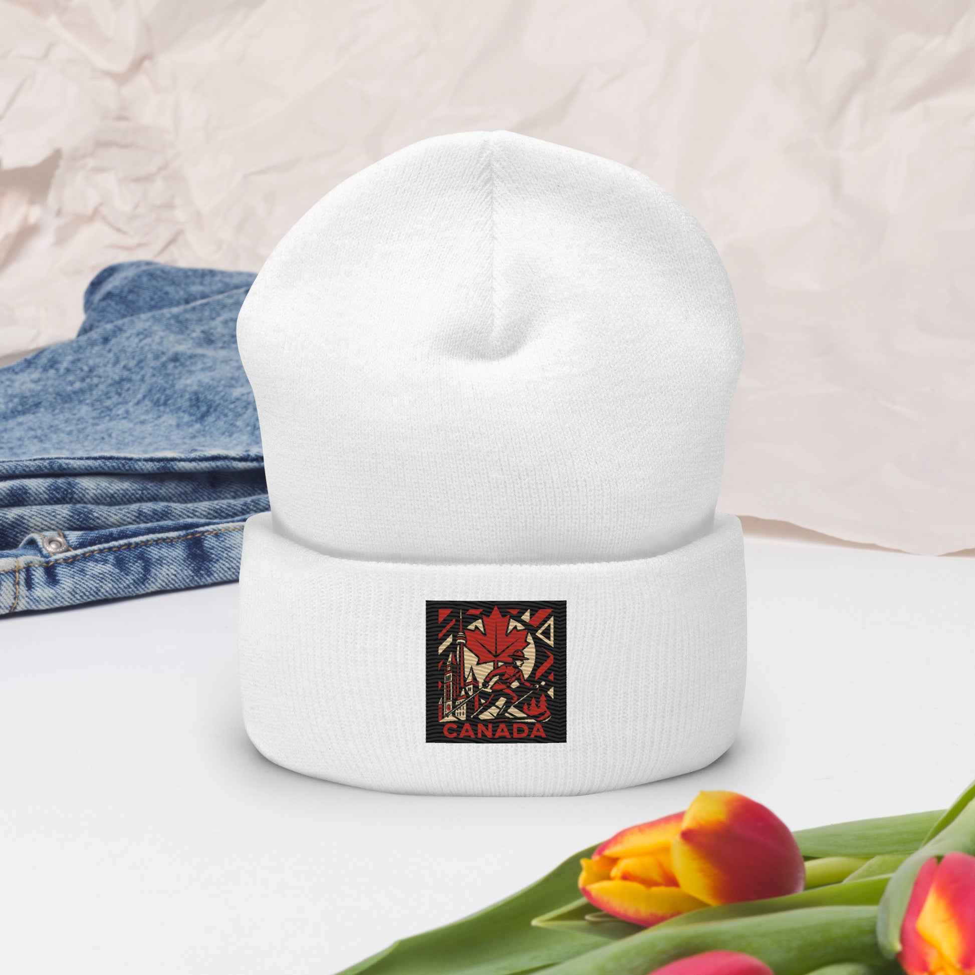 White beanie with a logo and text, placed on a surface with tulips and denim jeans in the background.