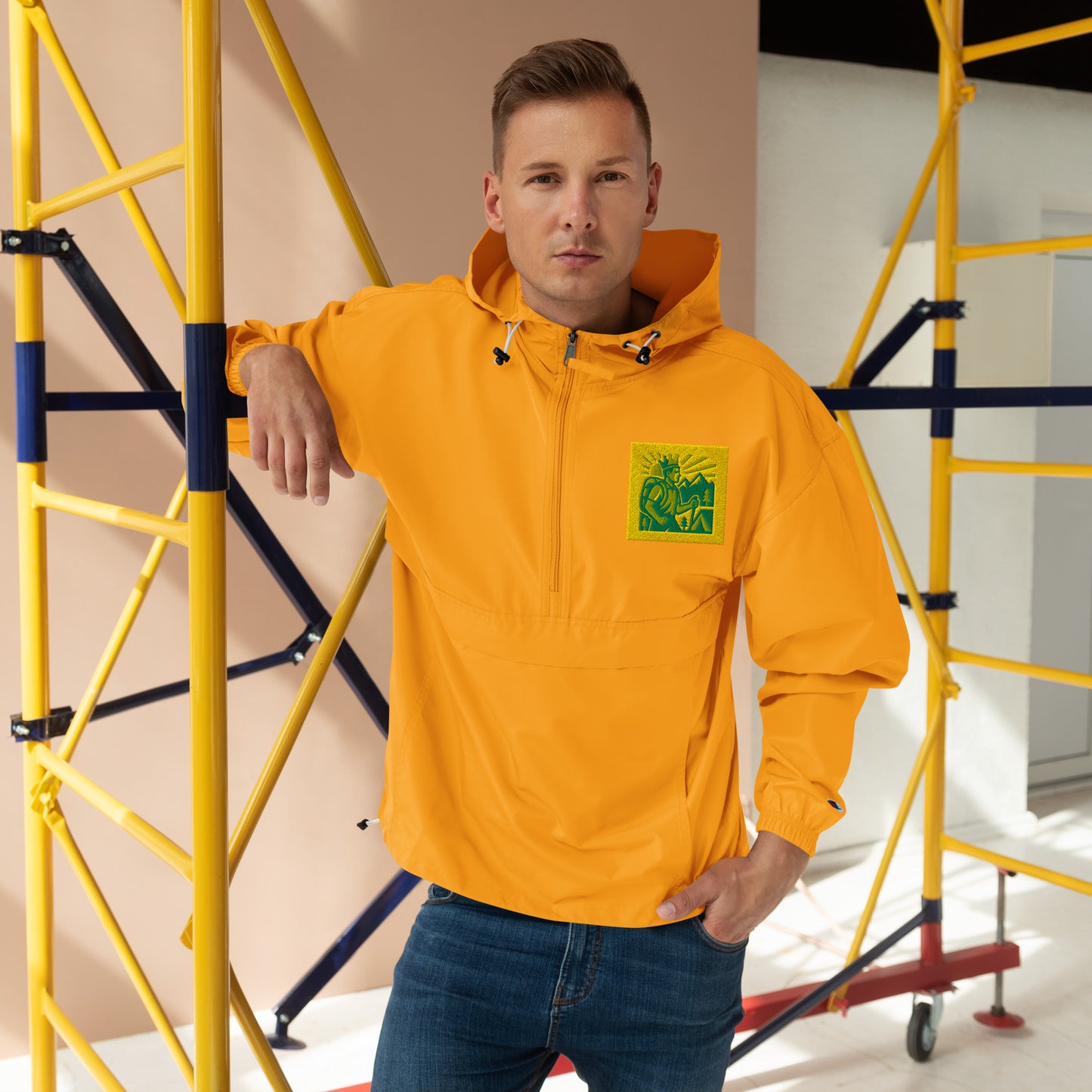 Man wearing a bright orange jacket with a green logo, standing in front of yellow scaffolding.