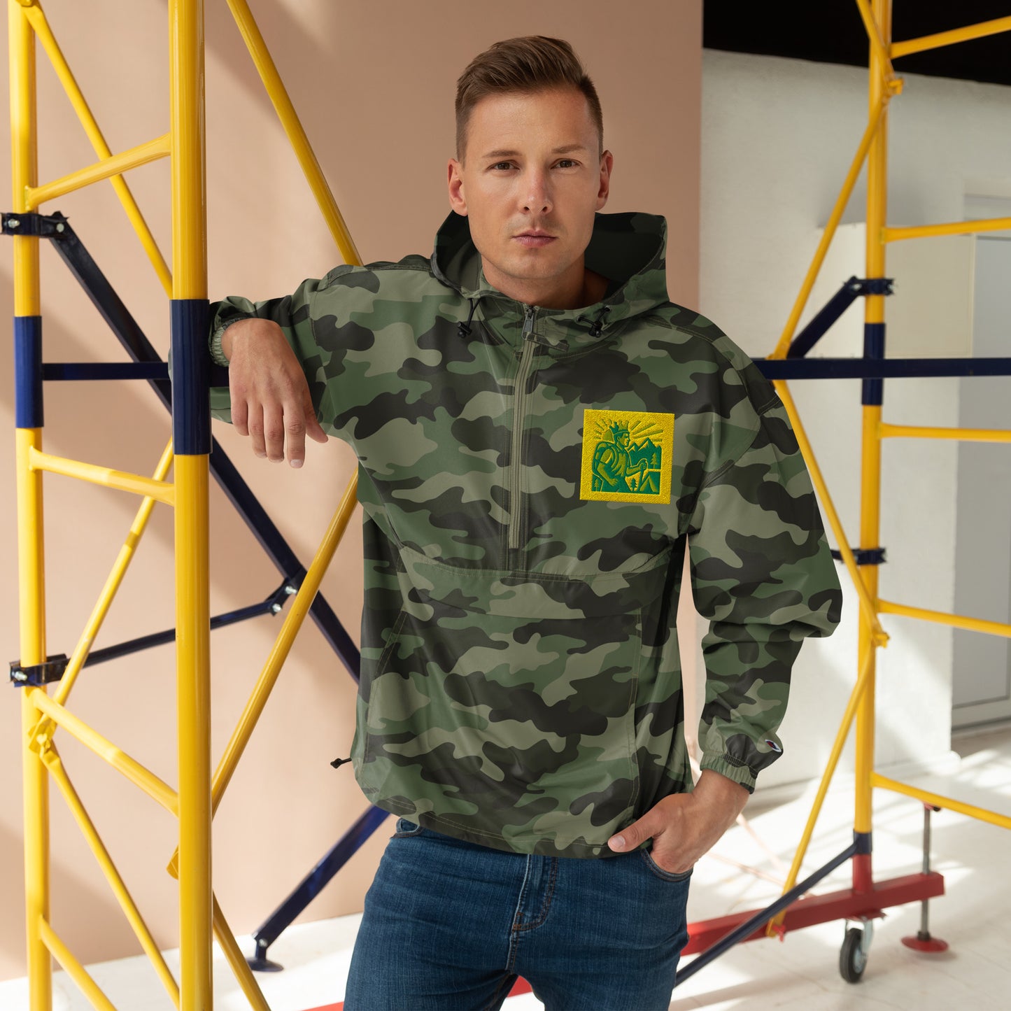 Man wearing a camouflage jacket with a logo, standing in front of scaffolding.
